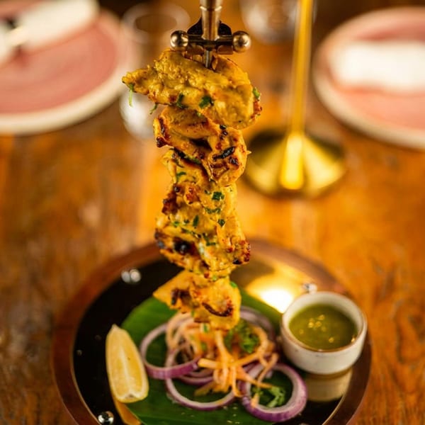 Skewer of grilled chicken tikka with onions, lemon, and green chutney on a wooden table.