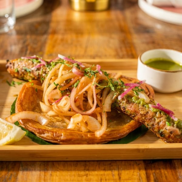 Grilled kebabs with sliced onions, herbs, lemon, and green chutney on a wooden tray.