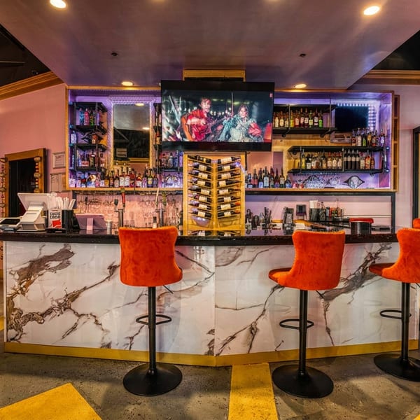 Modern bar with marble counter, orange stools, liquor bottles, and a TV screen.